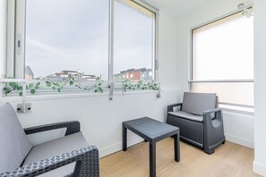 Ein Balkon mit Rattanmöbeln, einem kleinen Tisch und Blick auf Gebäude durch große Fenster.