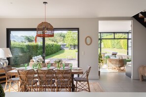 Comedor con mesa lista para seis personas, sillas de mimbre, lámpara colgante y vista a un jardín a través de ventanas amplias.