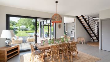 Une salle à manger spacieuse, avec une grande table dressée pour six personnes, entourée de chaises en osier, d’un luminaire suspendu et offrant une vue sur un jardin grâce à de grandes portes vitrées.