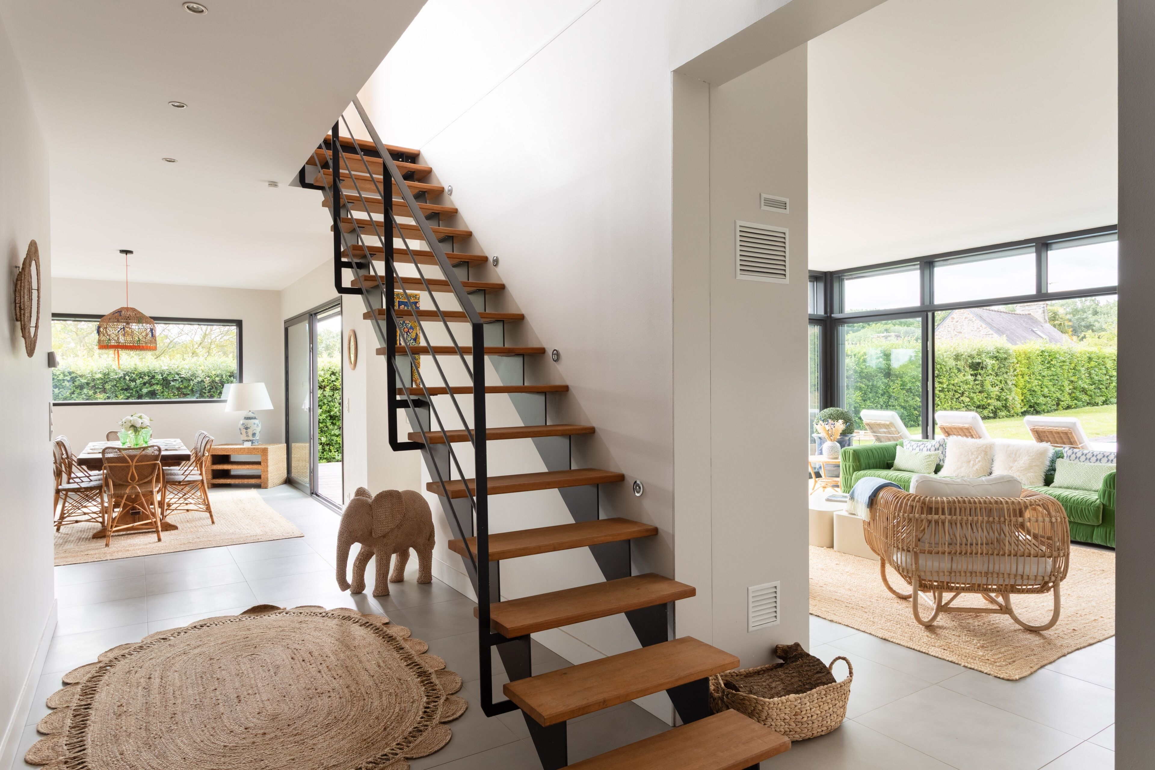 Un intérieur moderne avec un escalier en bois, un coin repas avec des meubles en osier et un canapé vert dans une pièce lumineuse dotée de grandes fenêtres.