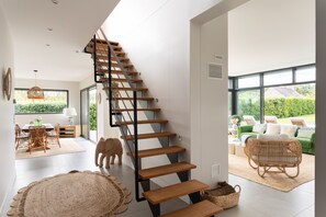 Ein modernes Interieur mit einer Holztreppe, einem Essbereich mit Korbmöbeln und einem grünen Sofa in einem hellen Raum mit großen Fenstern.