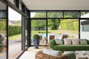 Ein modernes Interieur mit großen Fenstern, einem grünen Sofa, Korbmöbeln und Blick auf einen Garten.