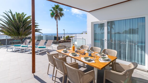 Zona pranzo con tavolo apparecchiato per sei persone, circondata da sedie in vimini, con vista su piscina e palme.