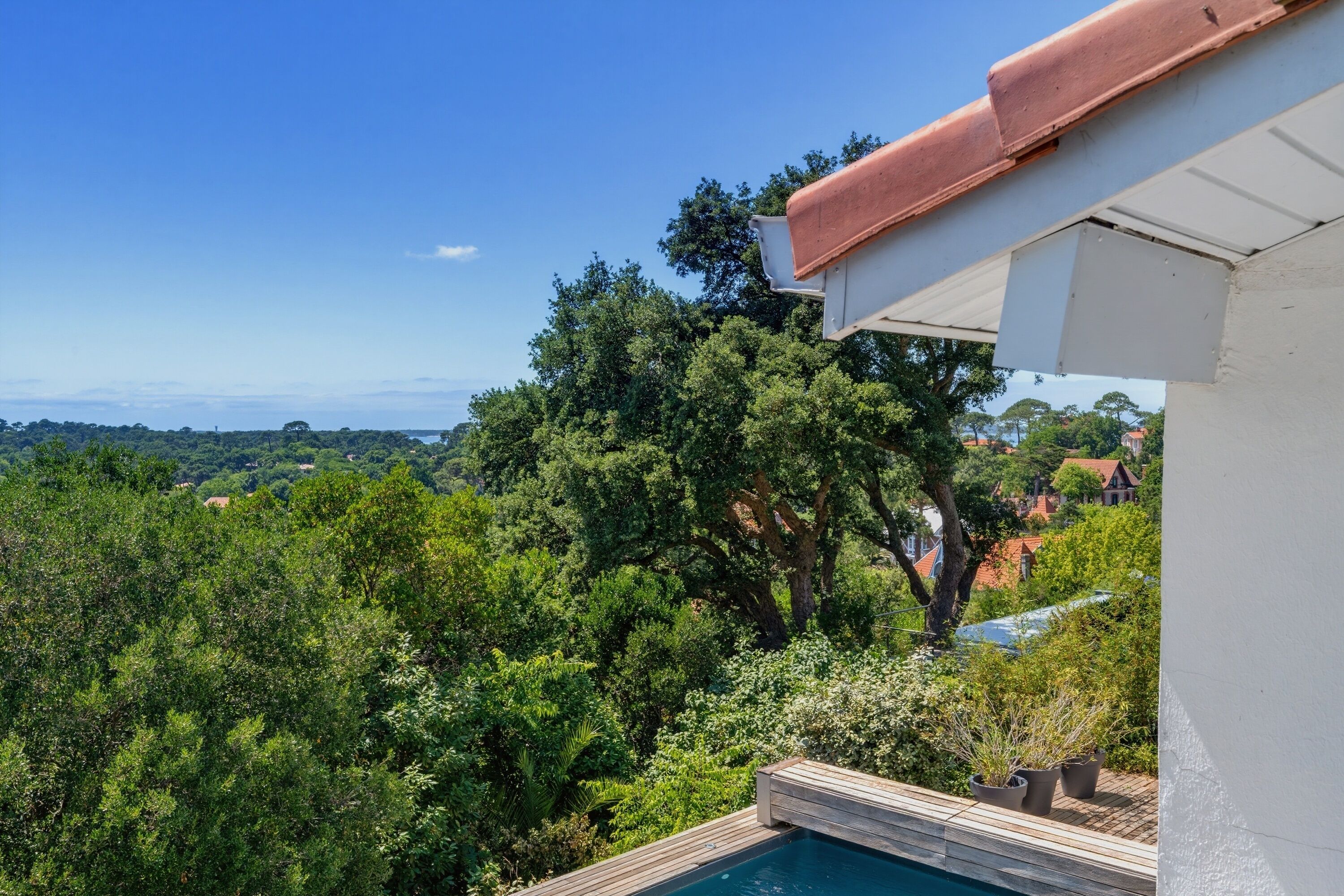 A pool with a wooden deck, surrounded by lush greenery and trees, with a view of distant houses and a clear blue sky.