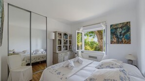 A bedroom with a large mirror, a bedside table with a lamp, a window with a view of greenery, and a painting on the wall. - La Vigie - Ville d'Hiver (Arcachon)