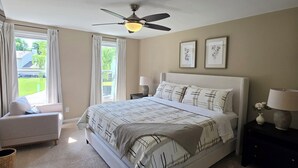 Un dormitorio con cama, ventilador de techo, mesita de noche con lámpara, ventana con cortinas e imagen enmarcada en la pared.