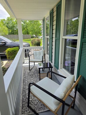 A covered porch with a white railing, a black metal chair, a small table, and a view of a green fence and trees. - Spacious Colonial-style single-family home (Waldorf)