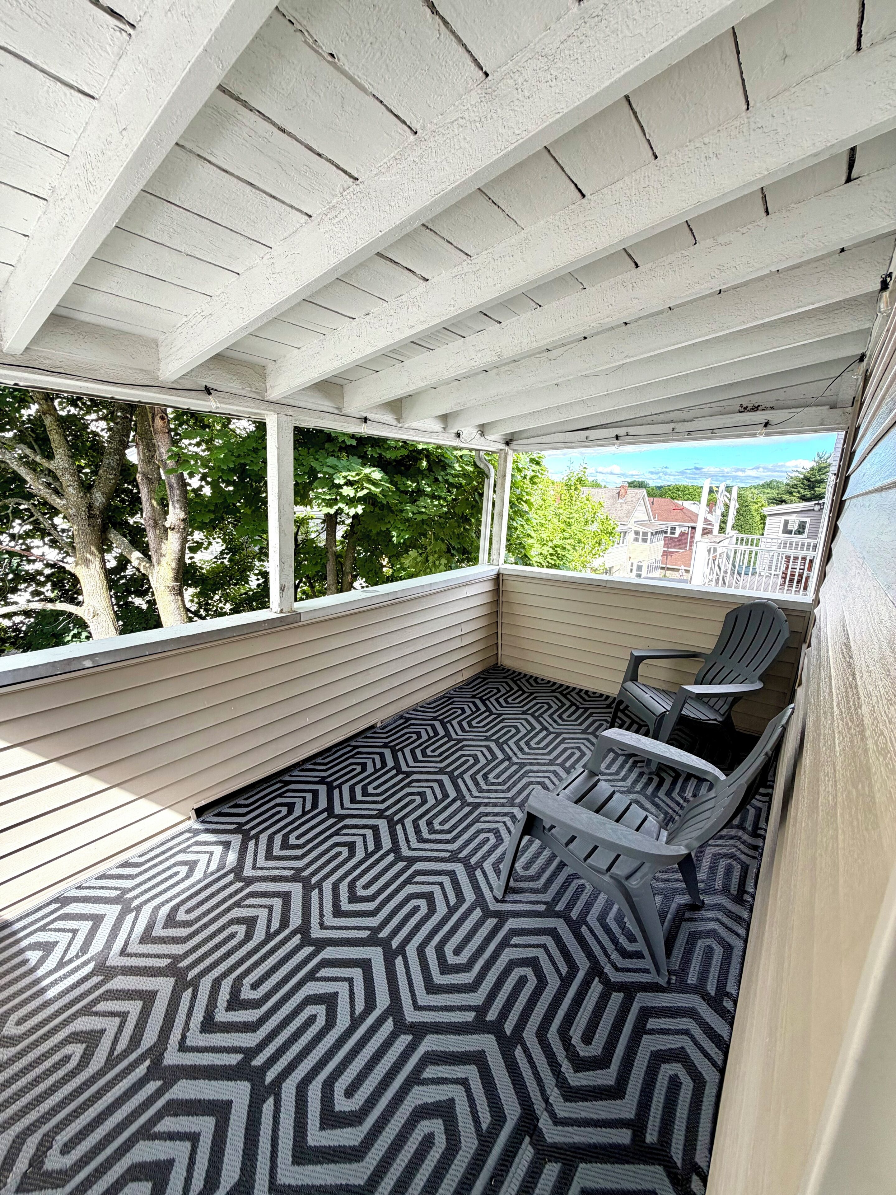 A covered outdoor seating area with a patterned floor, white walls, and a view of buildings and trees.