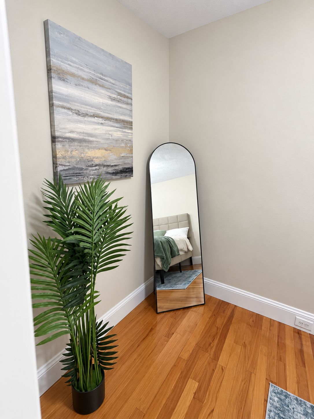 A room with a wooden floor, a large mirror, a potted plant, and a painting on the wall.