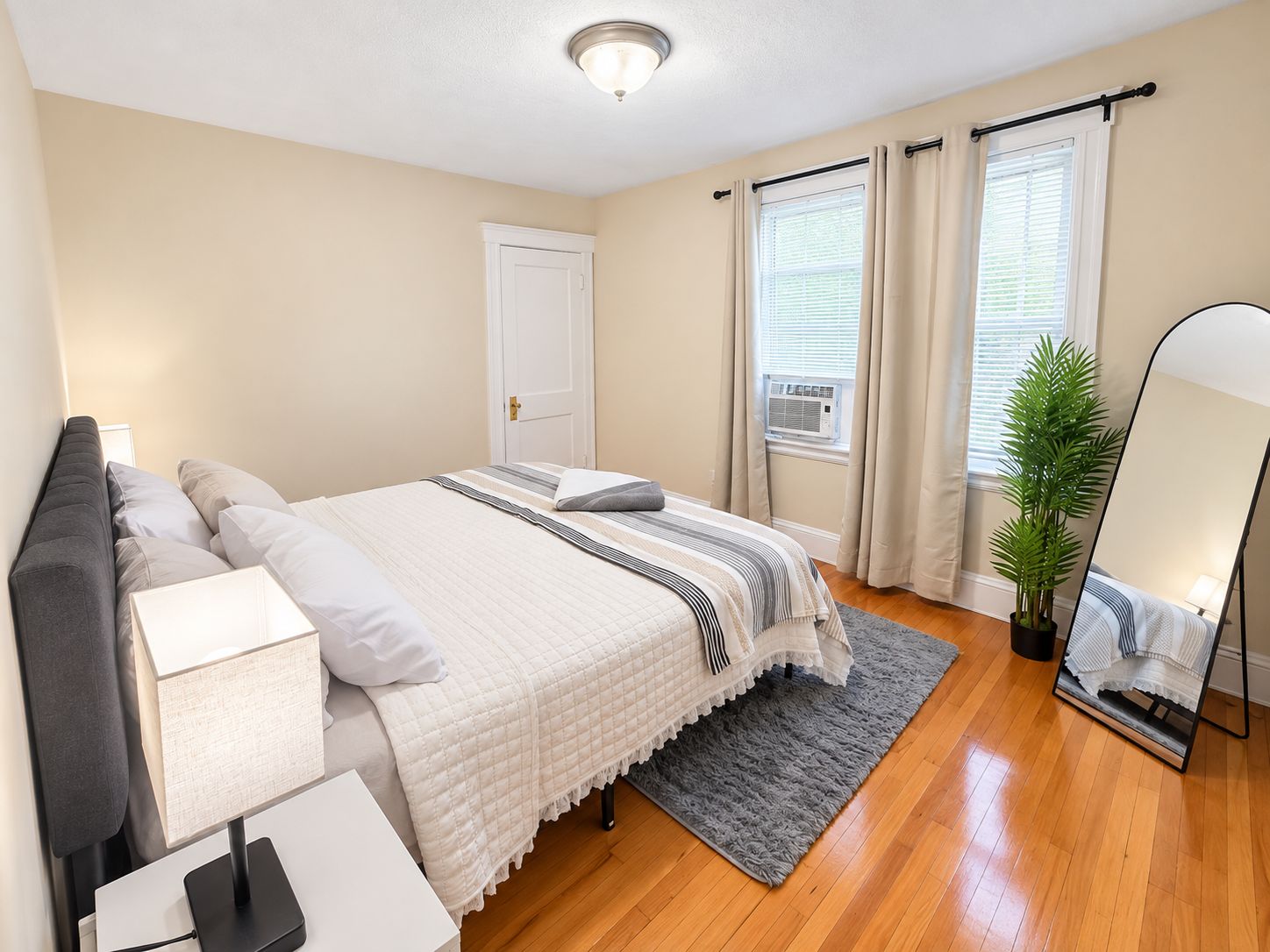 A bedroom with a bed, bedside table, lamp, window with curtains, and a potted plant.