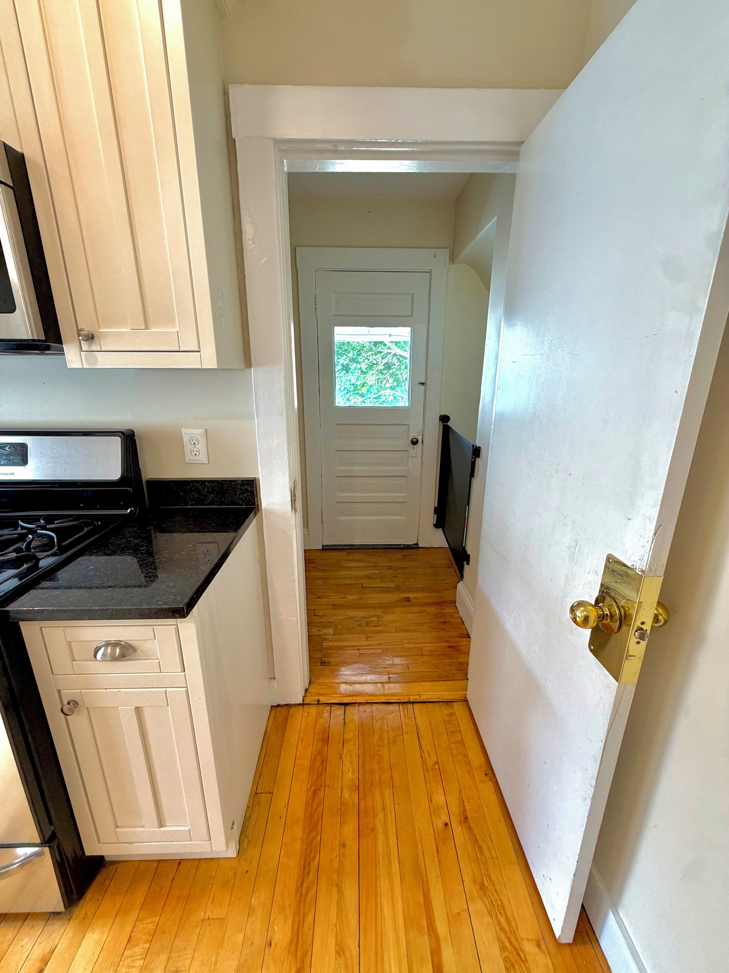 A narrow hallway with wooden flooring, a white door with a window, and a kitchen area with white cabinets and a black countertop.