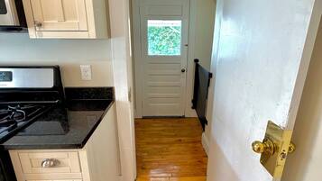 A narrow hallway with wooden flooring, a white door with a window, and a kitchen area with white cabinets and a black countertop.