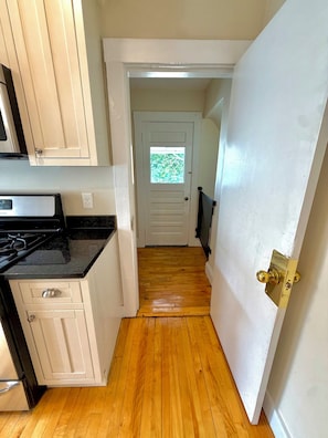 A narrow hallway with wooden flooring, a white door with a window, and a kitchen area with white cabinets and a black countertop. - 5AC Driveway
4BR
Tufts ~ Downtown Boston (Somerville)
