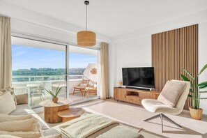 A modern living room with a large TV, comfortable seating, a balcony with a view, and a wooden accent wall. - Oceana Gardens Apartment (Cancelada)