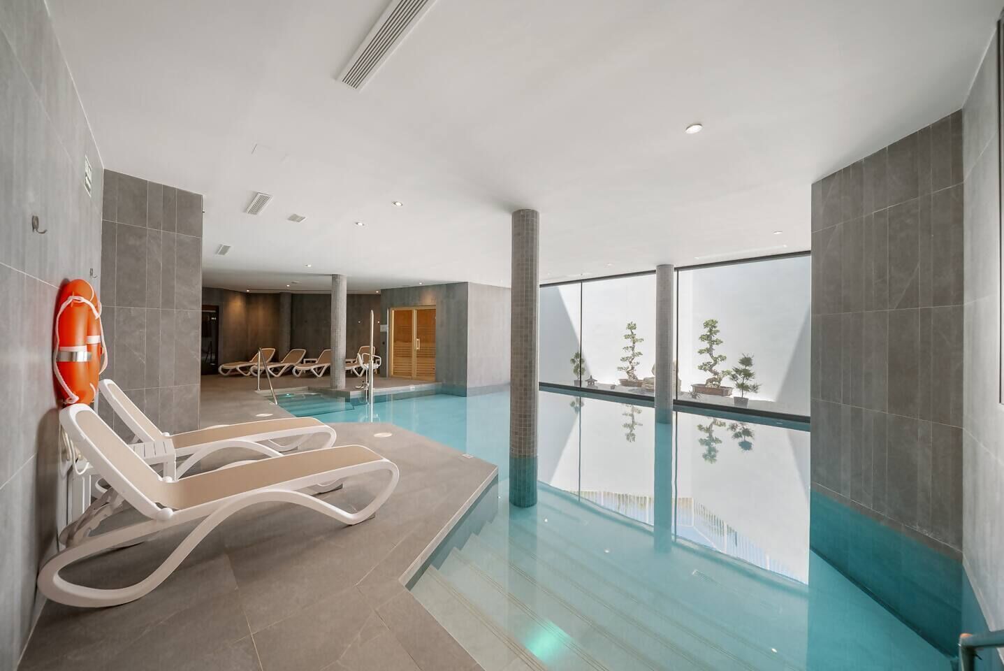 Una piscina interior moderna con paredes de vidrio transparente, sillas de descanso y un flotador en la pared.