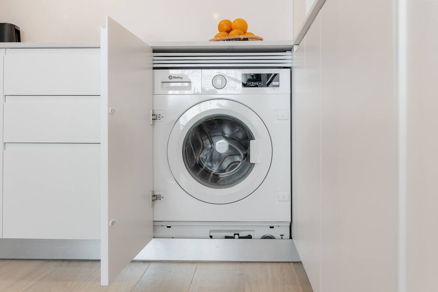 Un lave-linge compact et moderne, installé dans un petit espace buanderie, avec un meuble blanc et un panier rempli d’oranges posé sur le dessus.