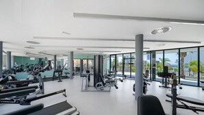Ett modernt gym med konditionsmaskiner, styrketräningsutrustning och stora fönster med utsikt över palmer och en klar himmel.