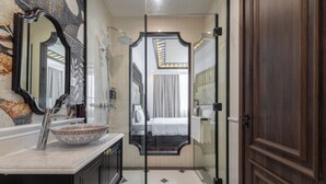A bathroom with a large mirror, a sink with a decorative bowl, a shower area, and a door leading to a bedroom.