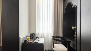 A compact hotel room with a small desk, a chair, a mirror, and a window with sheer curtains.