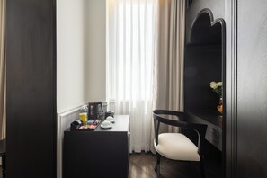 Ein kleines Hotelzimmer mit Schreibtisch, Stuhl, Spiegel und einem Fenster mit durchscheinenden Vorhängen.