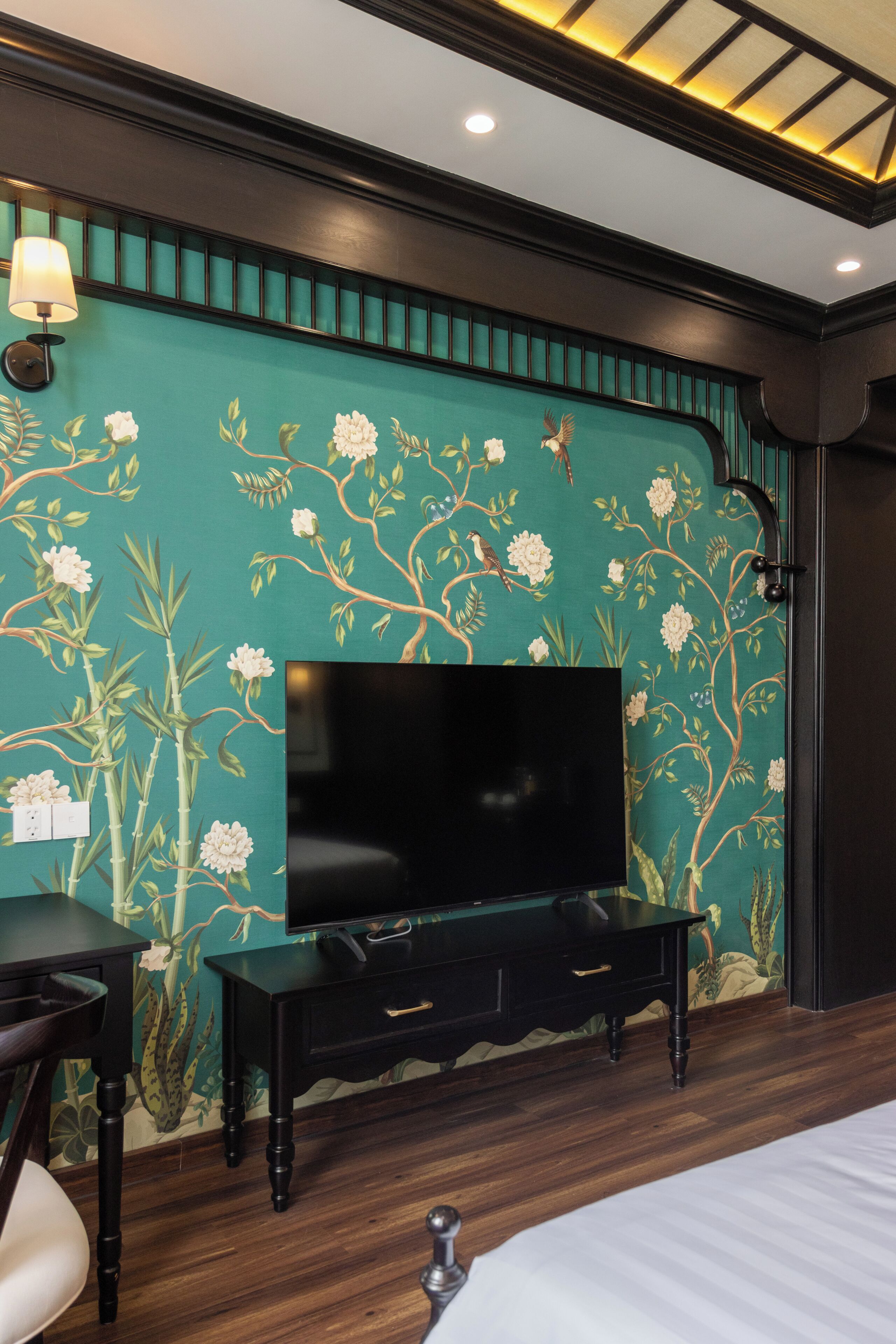 Una habitación con una televisión sobre un soporte negro, una cama, un escritorio con una silla y una pared con estampado floral.