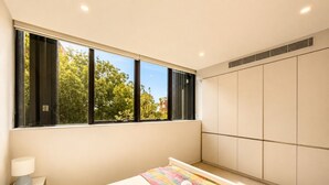Ein modernes Schlafzimmer mit großem Fenster, einem Bett mit bunter Decke, einer Nachttischlampe und einem Kleiderschrank.