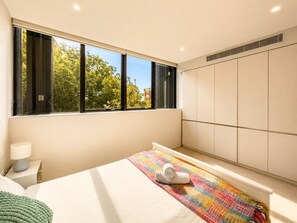 Ein modernes Schlafzimmer mit großem Fenster, einem Bett mit bunter Decke, einer Nachttischlampe und einem Kleiderschrank.