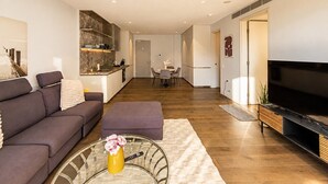 Ein modernes Wohnzimmer mit Sofa, Hocker und Couchtisch, ein Küchenbereich mit Esstisch und einem Flachbildfernseher.