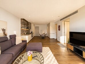 Ein modernes Wohnzimmer mit Sofa, Hocker und Couchtisch, ein Küchenbereich mit Esstisch und einem Flachbildfernseher.