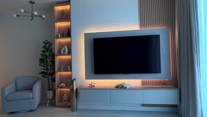 Un soggiorno moderno con una grande TV a schermo piatto montata su una parete bianca, una poltrona grigia, una pianta e uno scaffale con oggetti decorativi.