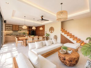 A modern living room with a sofa, dining table, and chairs, a staircase, and a kitchen in the background. - Villa Uma Atma Ubud by Nagisa Bali (Gianyar)