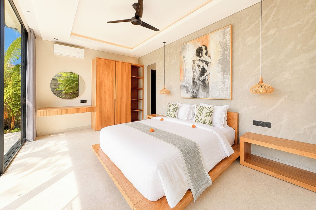 Un dormitorio moderno con una cama grande, un ventilador de techo, una obra de arte en la pared y vistas a zonas verdes al exterior.