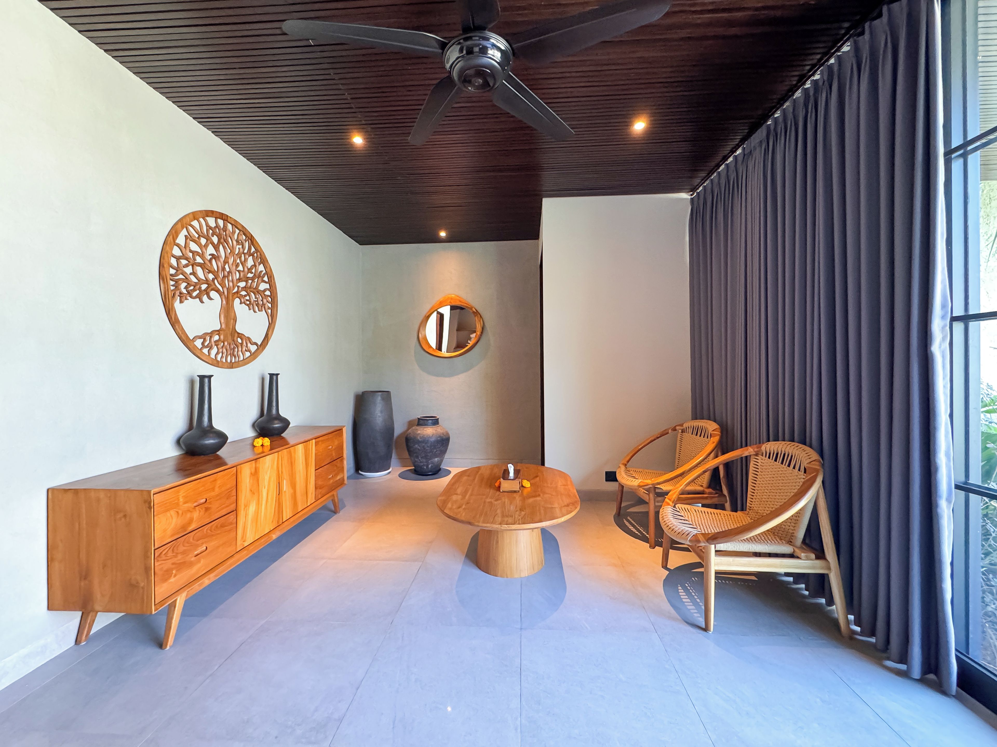 Un salon moderne avec un meuble en bois, une table basse ronde, un fauteuil en osier, un ventilateur de plafond et une grande fenêtre avec des rideaux.