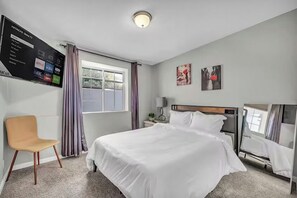 Ein Hotelzimmer mit einem Bett, einem Sessel, einem an der Wand befestigten Fernseher und einem Fenster mit Vorhängen.