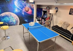 Ein Zimmer mit Tischtennisplatte, einer Couch, einer Spielkonsole und einer Wandmalerei zum Thema Weltraum.
