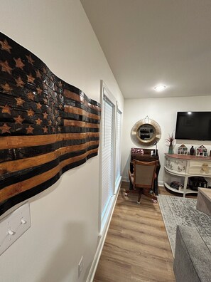 Una sala de estar con una decoración grande en la pared, un televisor de pantalla plana y un comedor con una silla y una mesa.