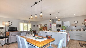 Una cucina moderna con mobili bianchi, un'area pranzo con tavolo di legno e sedie blu, e un soggiorno con divano e televisione.