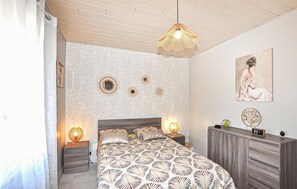 A bedroom with a patterned bedspread, a wooden headboard, bedside lamps, a wall clock, and decorative wall art. - Cozy home in Bagnoles (Bagnoles)