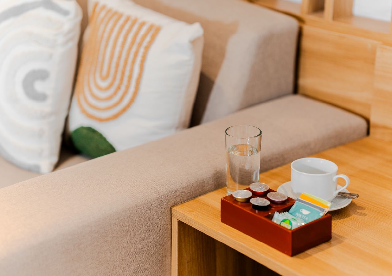 Una mesita de madera con un vaso de agua, una taza blanca y una bandeja con bolsitas de té y una caja de pañuelos.