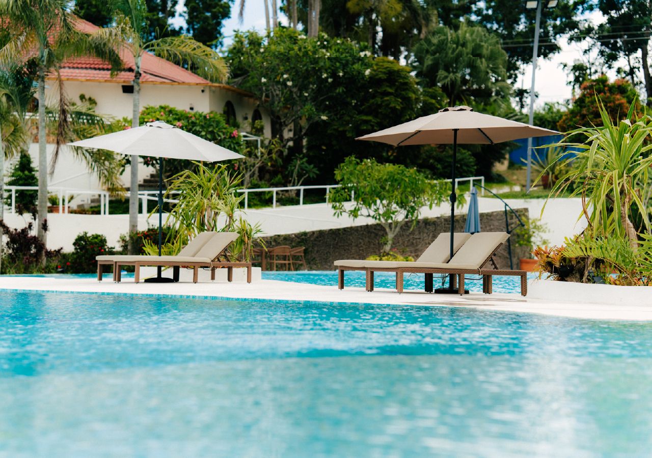 A swimming pool with lounge chairs and umbrellas, surrounded by greenery and a building with a red roof.