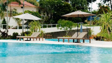 A swimming pool with lounge chairs and umbrellas, surrounded by greenery and a building with a red roof.