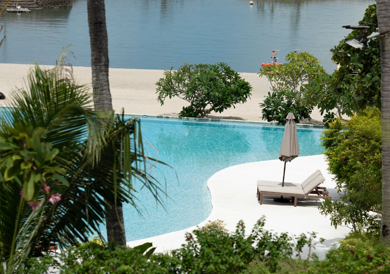 Piscina com deck branco, espreguiçadeiras e um guarda-sol.