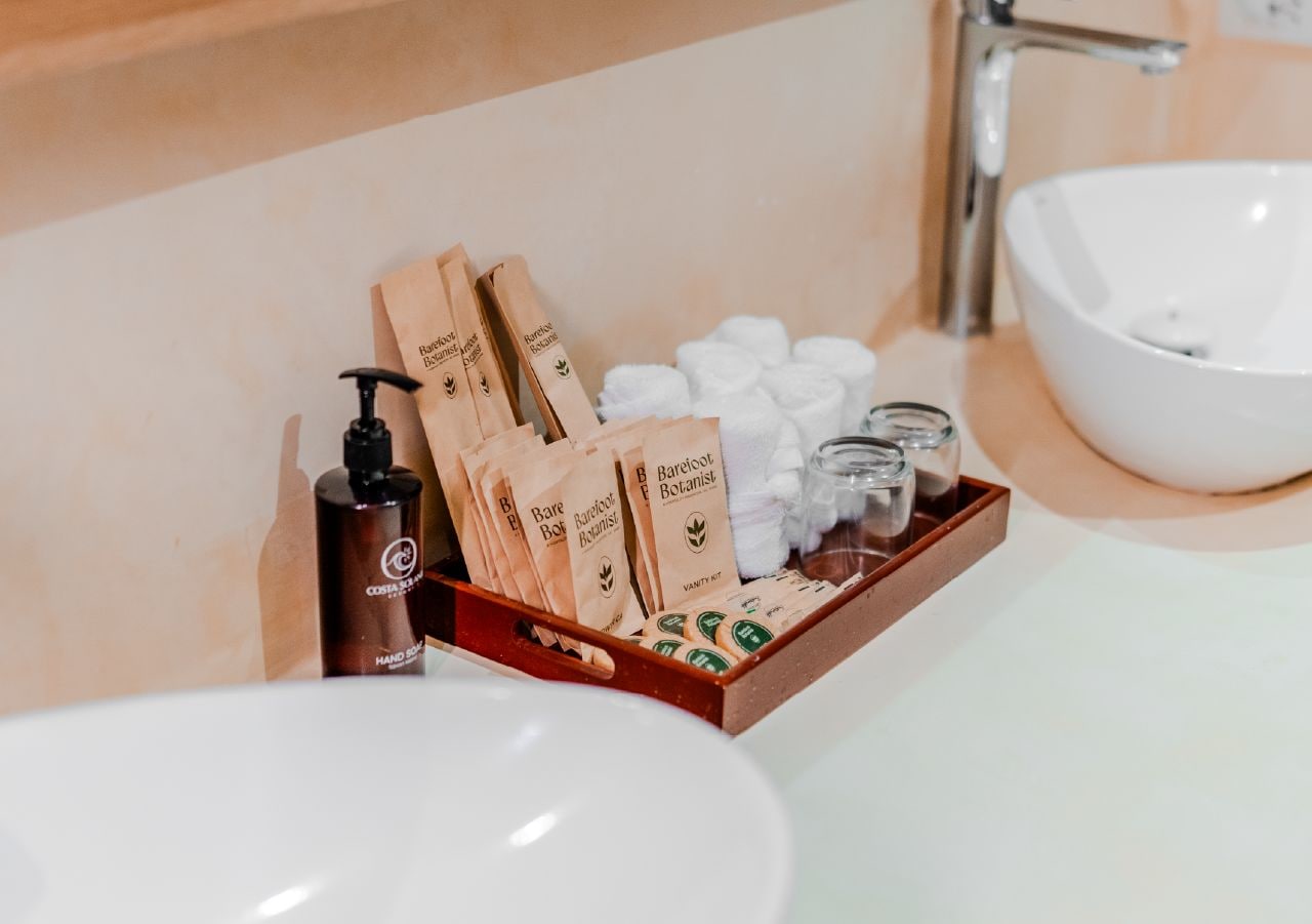 A bathroom vanity with a white sink, a bottle of hand soap, a wooden tray with toiletries, and a bottle of shampoo.