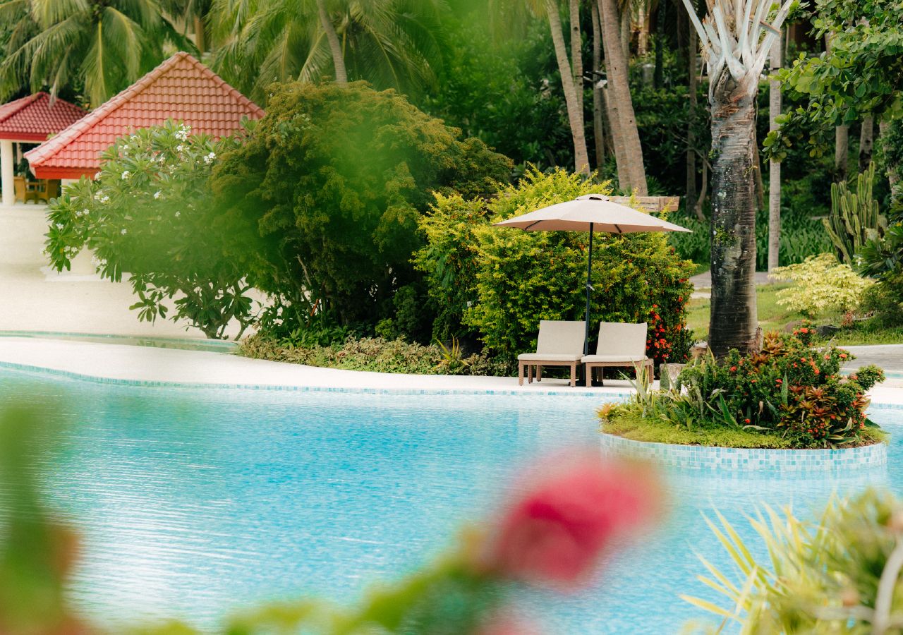 Un área de piscina con una silla de descanso y sombrilla, rodeada de follaje tropical y un pequeño edificio con techo rojo.