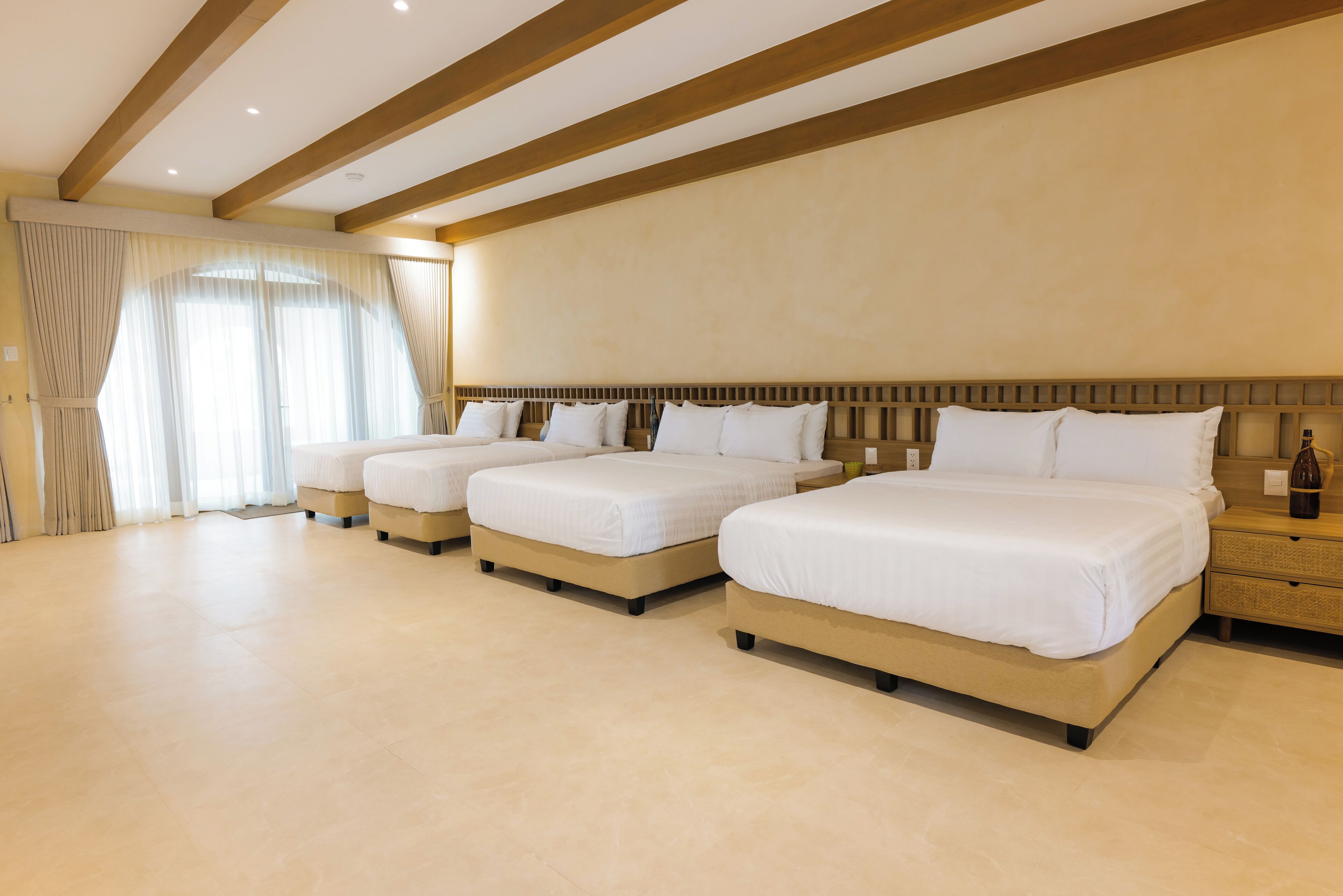 Habitación de hotel con tres camas, vigas de madera en el techo y un ventanal grande con cortinas.