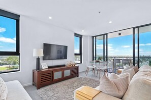 Ein modernes Wohnzimmer mit Flachbildfernseher, einer Couch, einem Esstisch mit Stühlen und großen Fenstern mit Blick auf die Stadt.