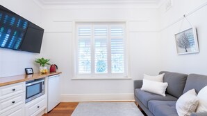 A modern living room with a grey sofa, white cushions, a flat-screen TV, a window with blinds, and a framed picture on the wall.