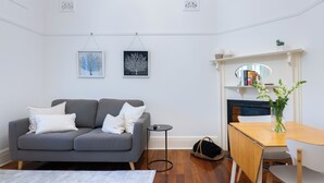 A living room with a grey sofa, a small round table, a wooden dining table, and a fireplace with a mirror above it.