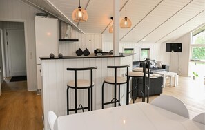 Una cuina moderna amb armaris blancs, una illa central amb tamborets, una zona de menjador amb taula blanca, i un saló amb sofà i televisió.