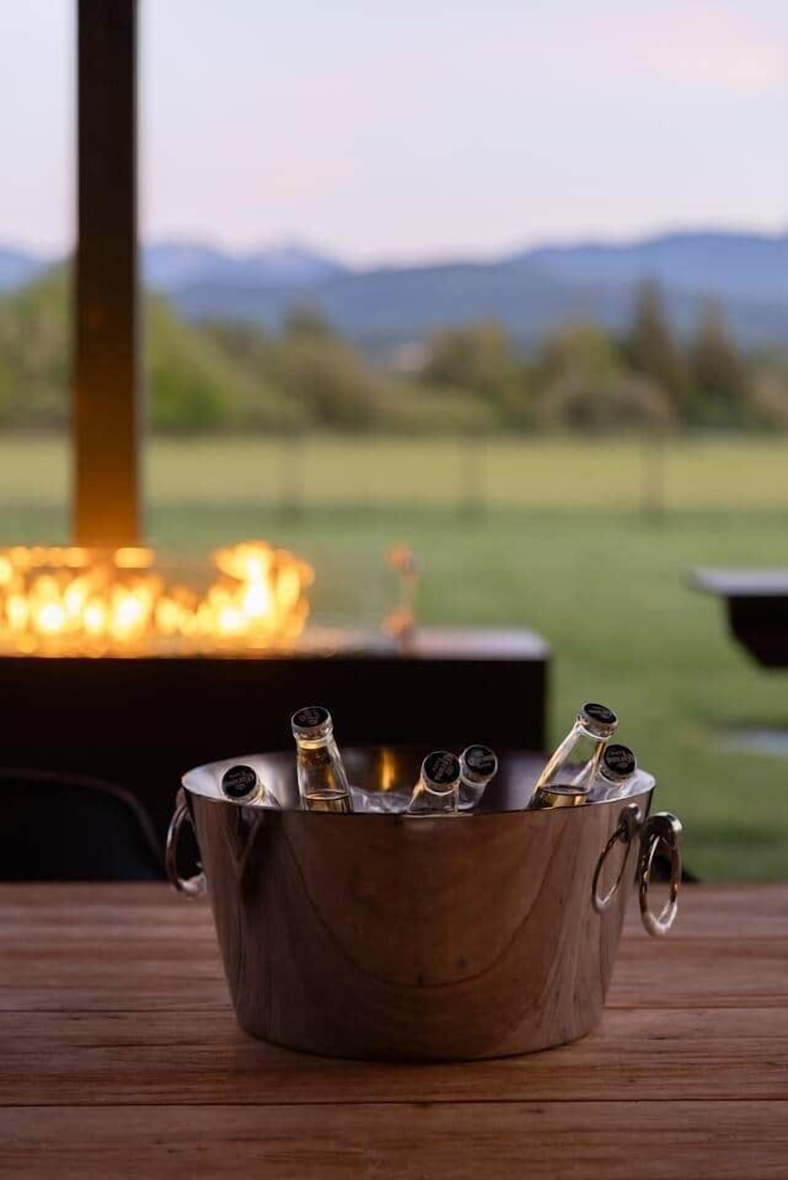 Ein Metallkübel mit Flaschen auf einem Holztisch, mit Feuer und Blick auf ein grasbewachsenes Feld und Berge im Hintergrund.